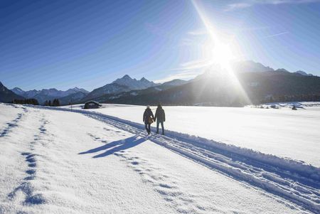 Zwei personen spazieren im Winter auf einem Weg durch die Zugspitz Region bei Sonnenschein und blauen Himmel.