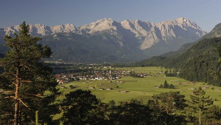 Von eine Anhöhe blickt man über den Ort Garmisch-Partenkirchen und grünen Wiese auf das felsige Wettersteinmassiv in der Zugspitz Region.