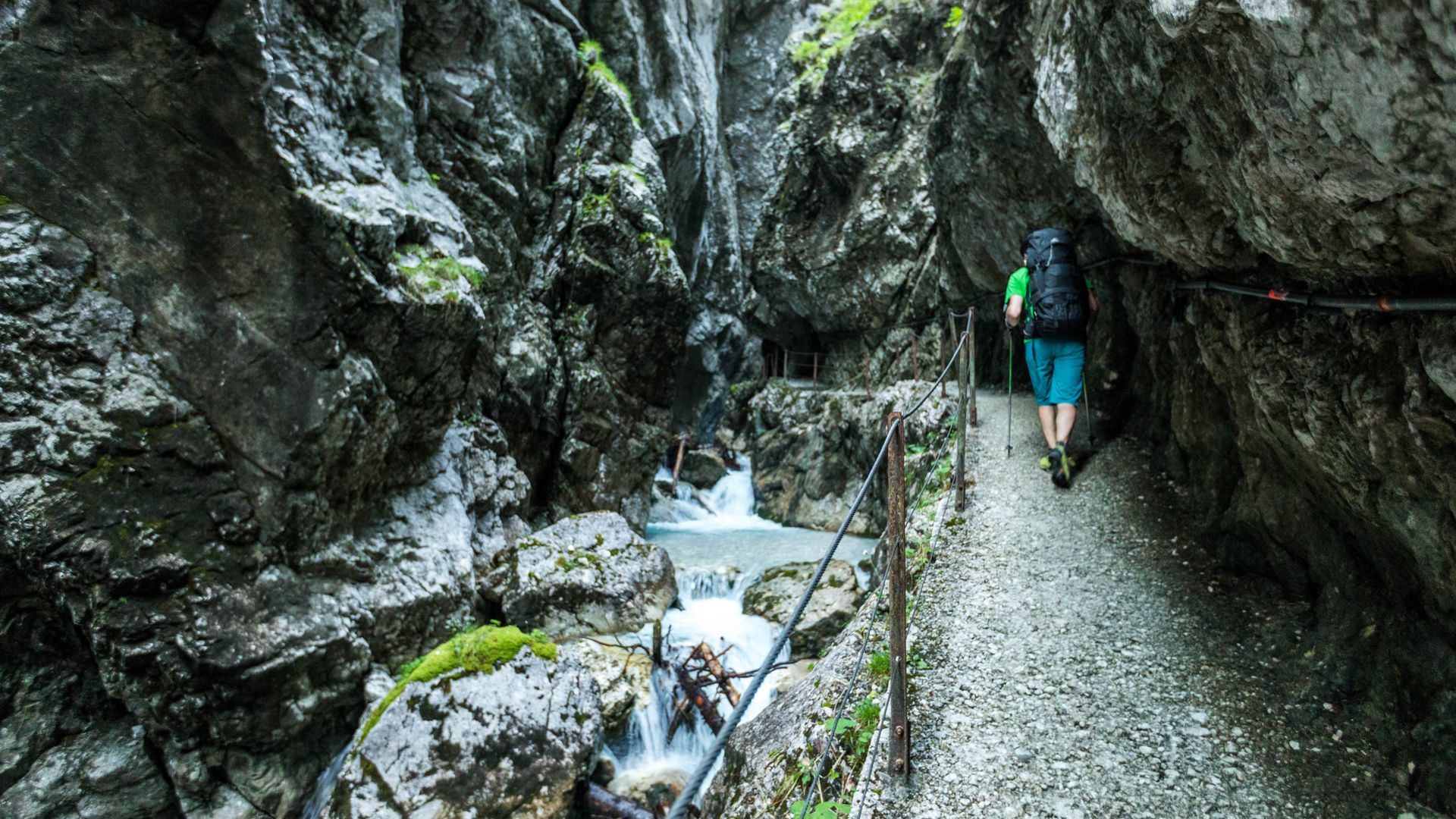 Rechts im Bild ist der Klammweg durch die Hööllentalklamm, eine Person geht gerade in einen Tunnel hinein. Links im Bild schlängelt sich der Bach durch die Felswände.