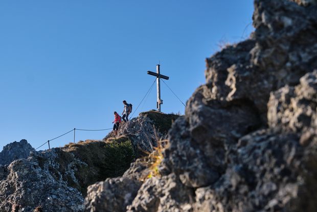Zwei Personen steigen von einem felsigen Gipfel mit Gipfelkreuz ab. Der Weg ist mit Drahtseil versichert.
