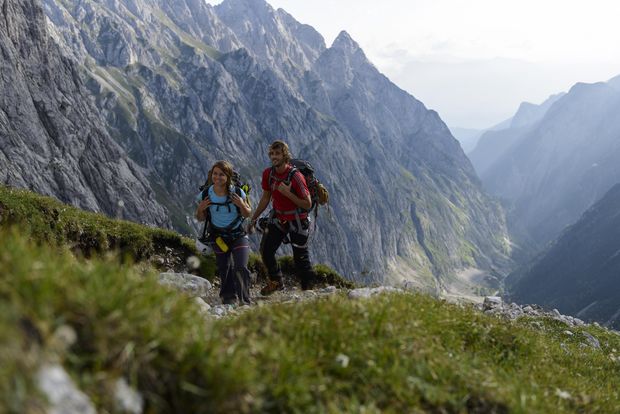 Zwei Personen gehen auf einem Wanderpfad zwischen einer Bergwiese bergauf. Im Hintergrund sieht man Berge und ein Tal.