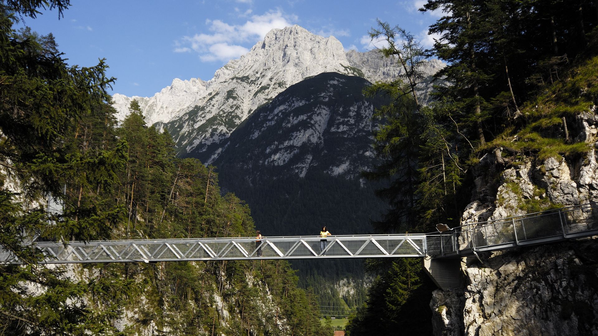Eine silberne Brücke führt über eine Schlucht, die Geisterklamm. Im Hintergrund ragt ein felsiger Berg empor.
