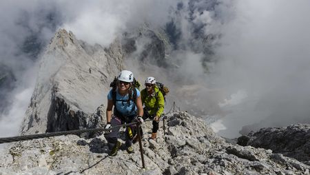 Zwei Personen mit Helm und Klettersteigausrüstung gehen auf einem felsigen Grat einen gesicherten Klettersteig.