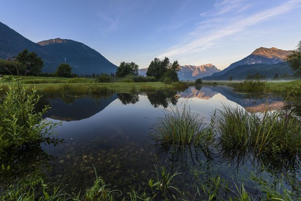 Im Vordergrund sieht man einen kleinen See mit viel grüne am Ufer. Im Hintergrund sieht man Berge, unter anderem das Wettersteingebirge, das sich im See spiegelt.